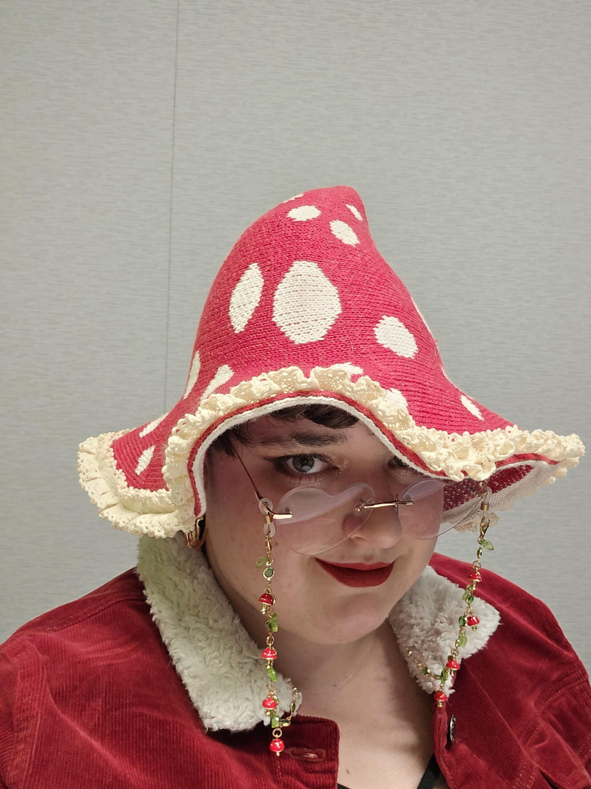 Person wearing a red polka-dotted hat with lace trim and a red coat, partial face visible with pink sunglasses and red lipstick amid beaded jewelry.