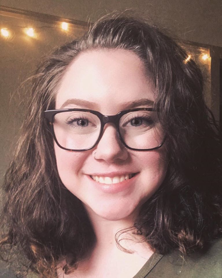 Young woman with dark wavy hair and black-framed glasses, smiling at the camera indoors with string lights in the background.