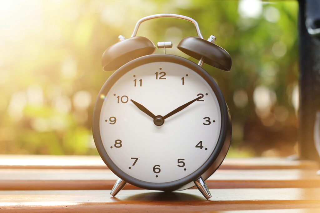 An alarm clock on a wooden table with greenery behind it.