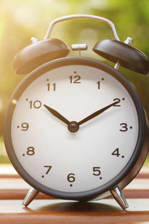 An alarm clock on a wooden table with greenery behind it.