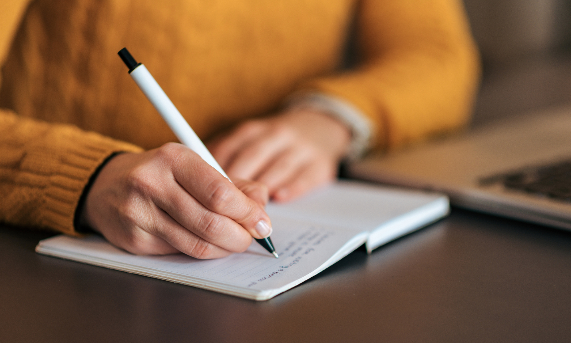 hands writing in a notebook, yellow sweater, white pen, notebook, laptop