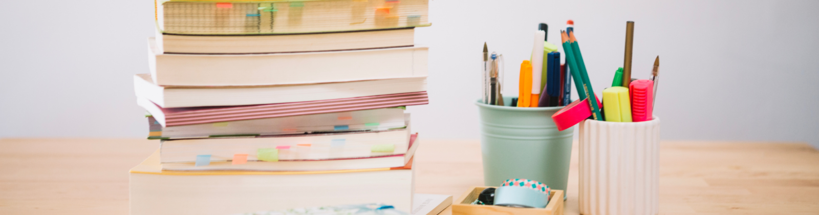 Stack of assorted books piled on a wooden desk beside a mint-colored container filled with colorful pens and pencils.